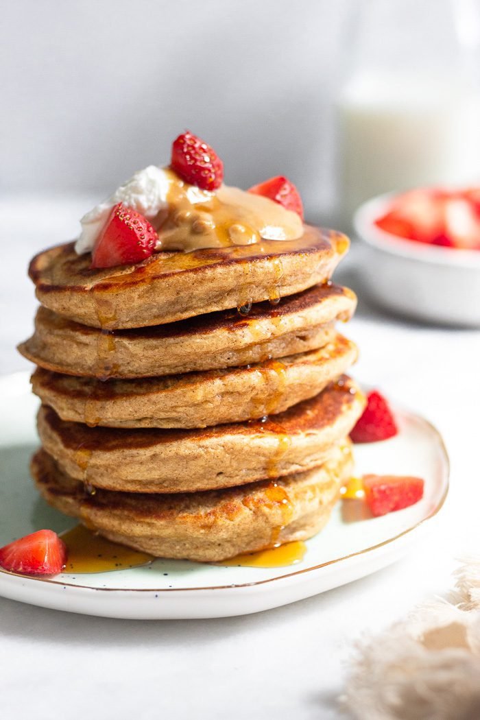 A stack of protein pancakes on a plate topped with yogurt, peanut butter, berries, and syrup.