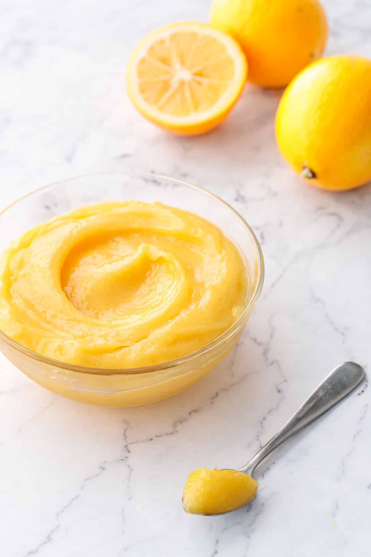 Glass bowl filled with Meyer Lemon Curd on a marble surface, with a spoonful of lemon curd in the foreground and a few fresh Meyer lemons out of focus in the background.