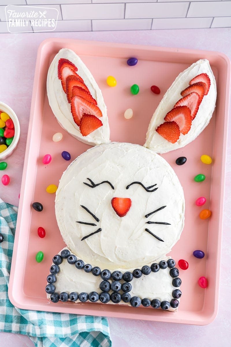 This bunny cake is so cute, that it’s almost an insult to eat it. But no worries, this Easter bunny cake is delicious as well as adorable! Find out how to make this fun cake at home. A bunny cake on a pink baking sheet decorated with strawberries, blueberries, and black icing in the shape of eyes and whiskers.