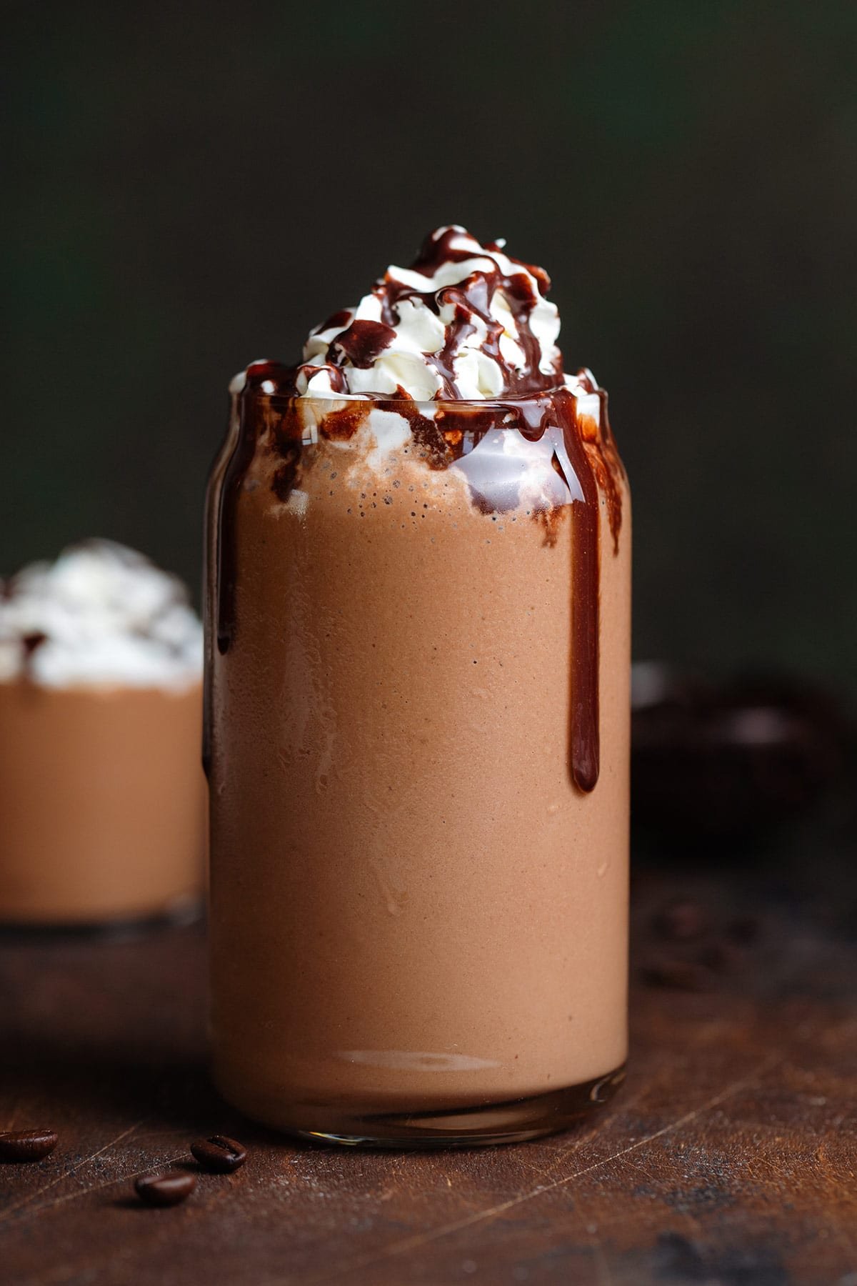 A dark brown mocha smoothie in a tall glass garnished with whipped cream and chocolate sauce on a dark wooden background.