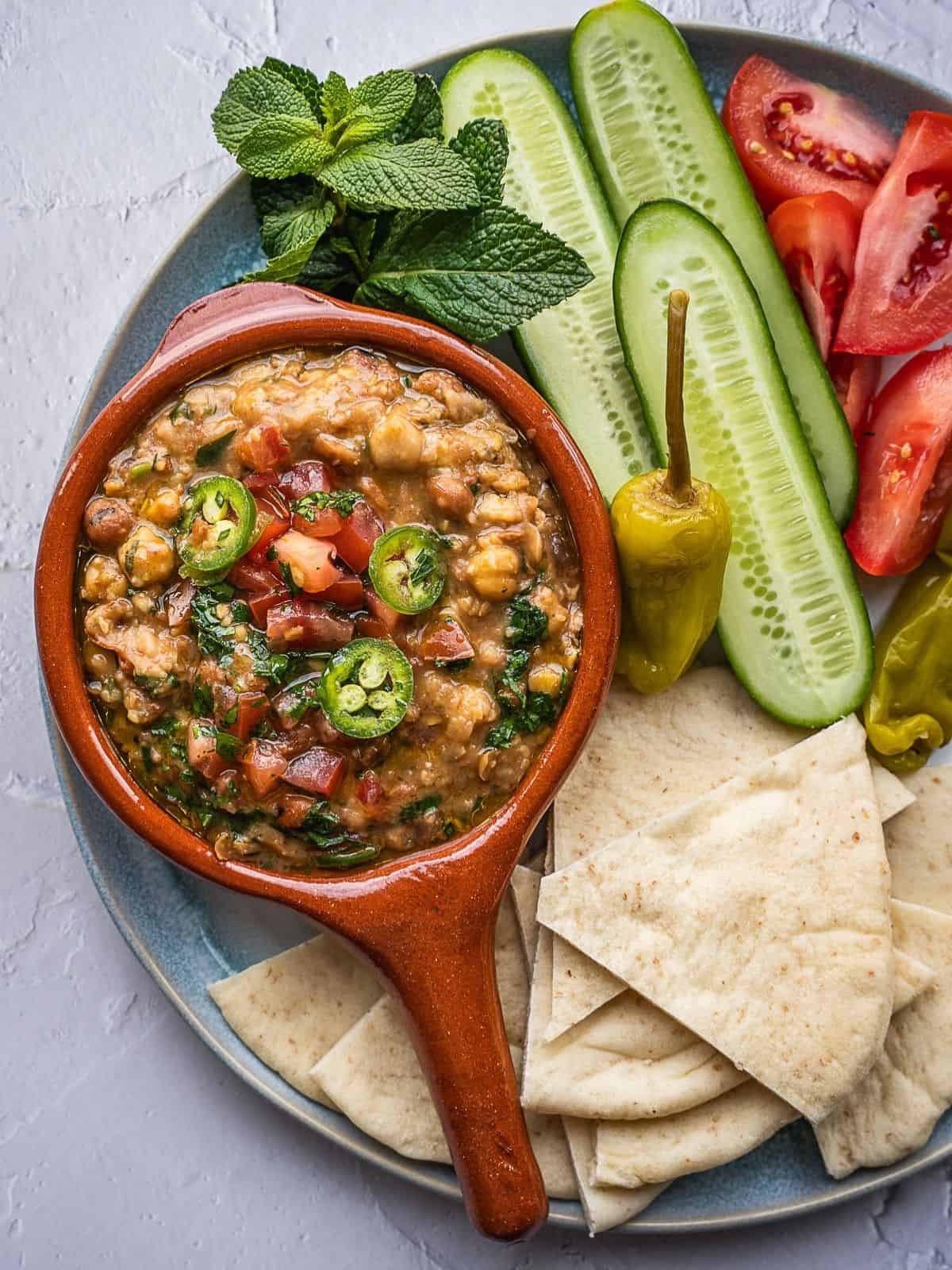 Platter with ful medames, sliced cucumbers, sliced tomatoes, and pita triangles. 