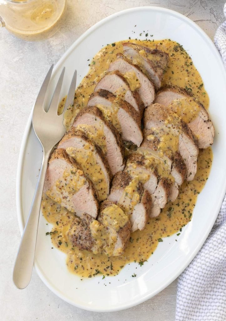 Slices of roast pork tenderloin with a creamy mustard sauce on a white platter.