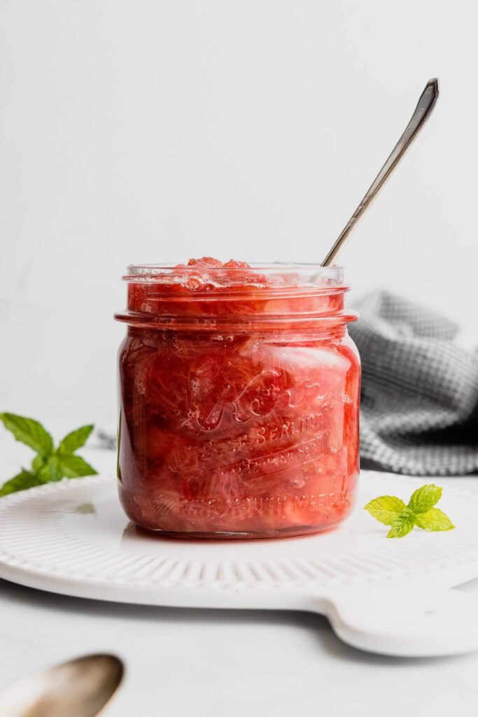 A glass jar filled with bright red rhubarb compote. There is a spoon in the sauce and the jar sits on a white tray. There's a mint sprig be the jar. 