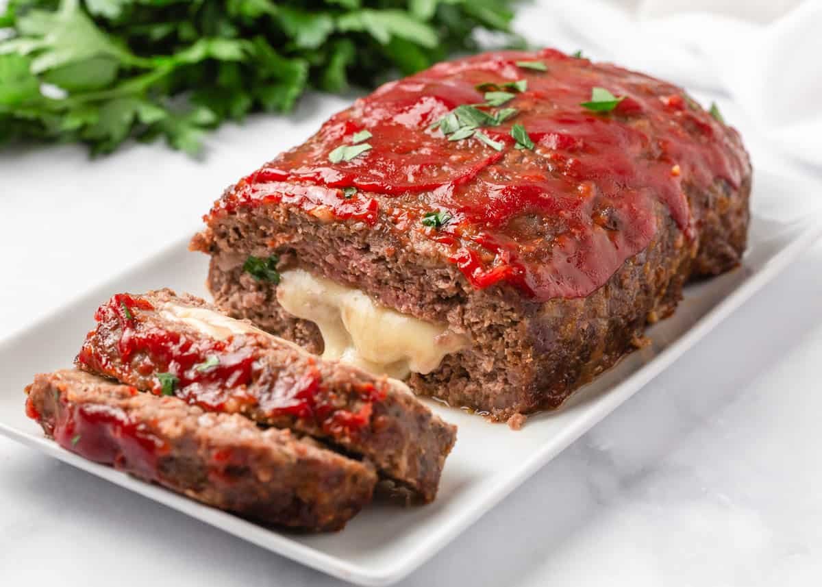We're adding a layer of cheese to a classic recipe making this cheesy mozzarella stuffed meatloaf an instant hit! Juicy seasoned meat, a melty middle, and a delicious sweet and tangy glaze on top makes this the ultimate comfort food! Sliced stuffed meatloaf on white plate.